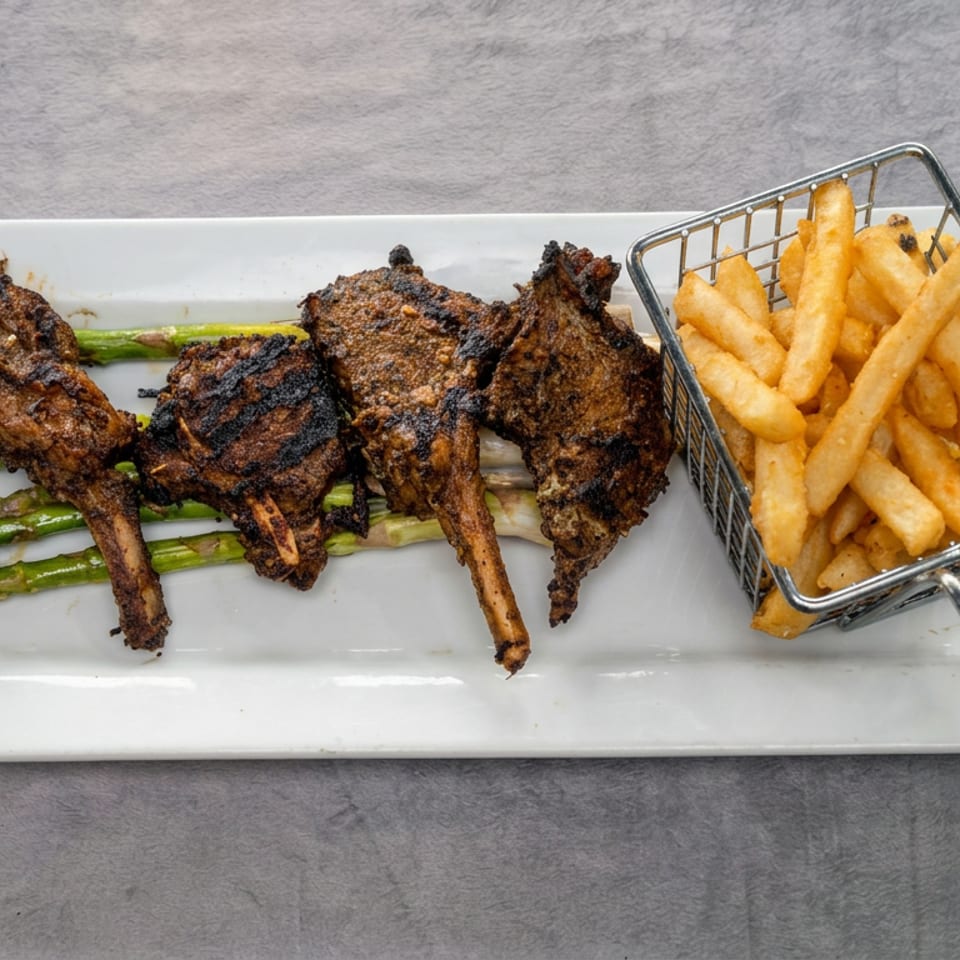 Grilled lamb chops served over asparagus with a basket of golden fries on a white rectangular plate.